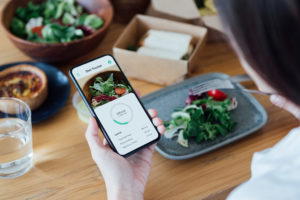 Over the shoulder shot of young woman using mobile app to track nutrition and count calories with smartphone while eating lunch.