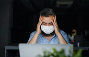 Frustrated mature man with face mask sitting indoors in office, working